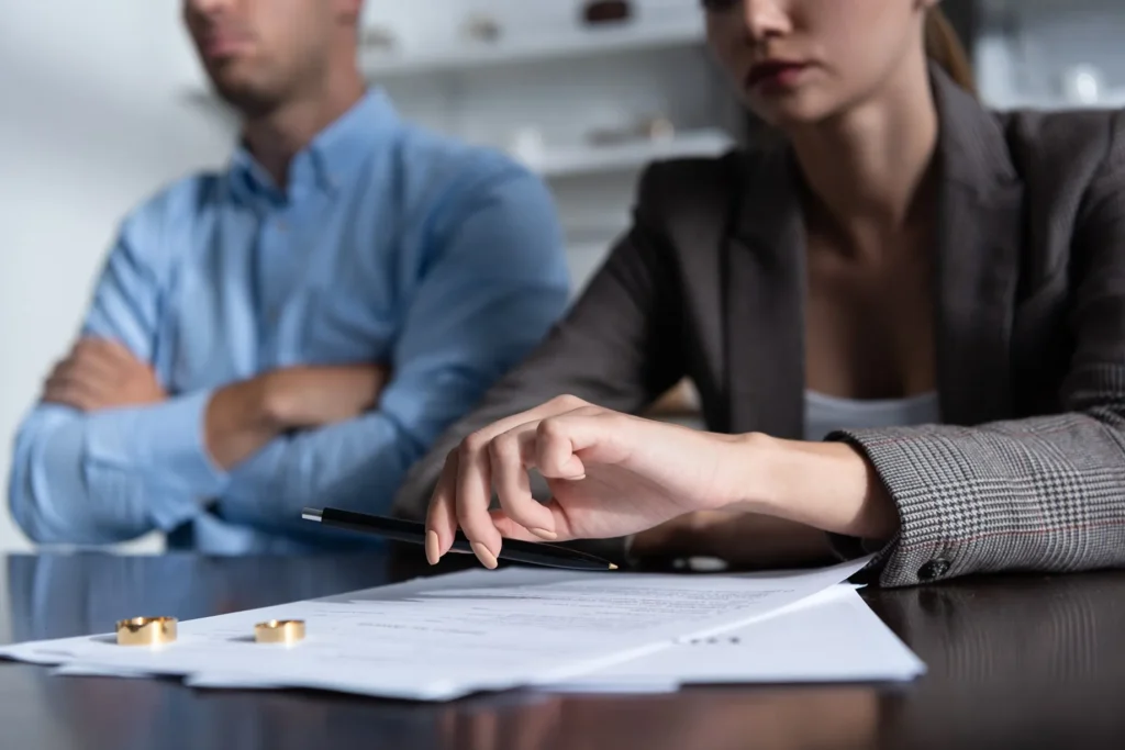 A woman picking up a pen to sign divorce papers with two wedding rings on the table with a man in the back.