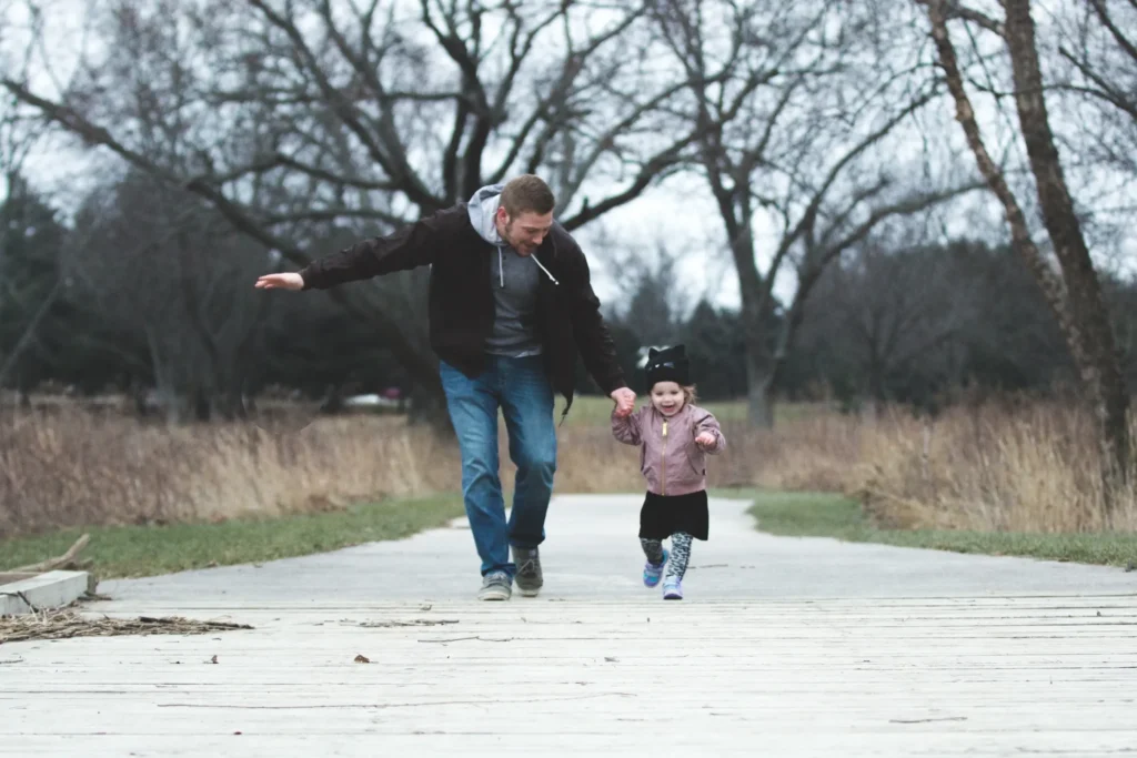 A man holding his daughter's hand walking down a sidewalk in the woods.