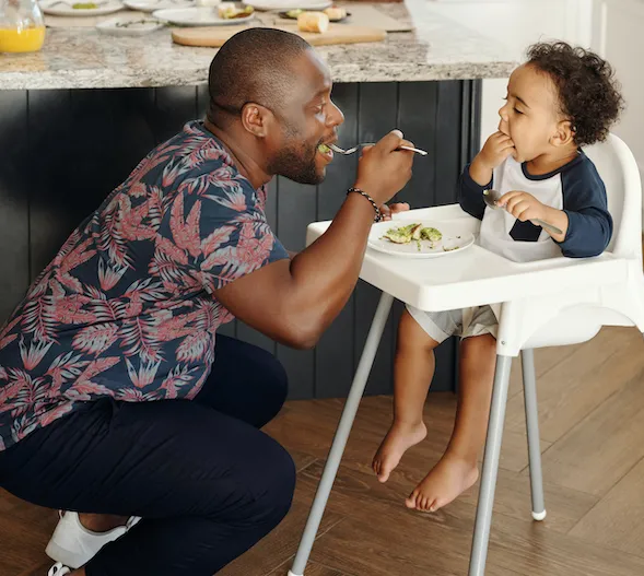 A baby eating in his high chair with his dad eating off his food plate with a fork.