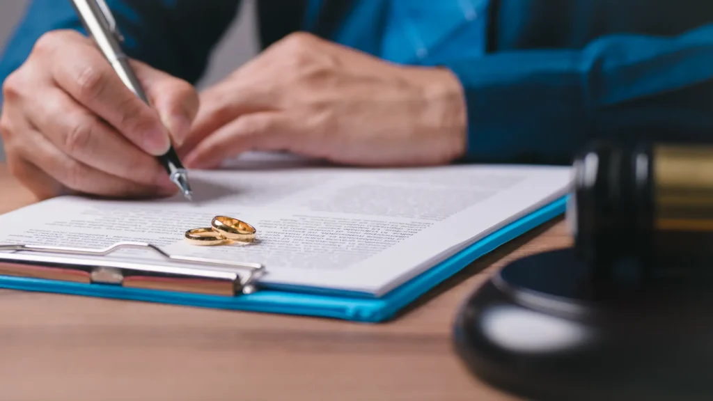A man signing papers with two gold rings and a gavel on the clipboard, symbolizing divorce proceedings.