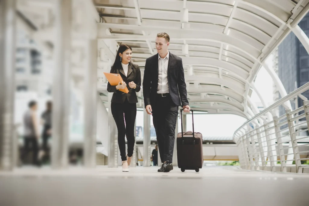 Two business professionals walking together in a modern urban walkway, one holding documents and the other pulling a suitcase, representing international business and legal services.
