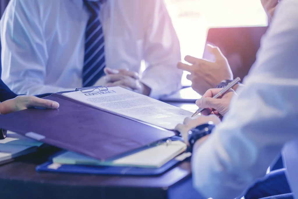 Group of business professionals sitting at a table reviewing and discussing a contract document on a clipboard, representing outside general counsel legal services for businesses.