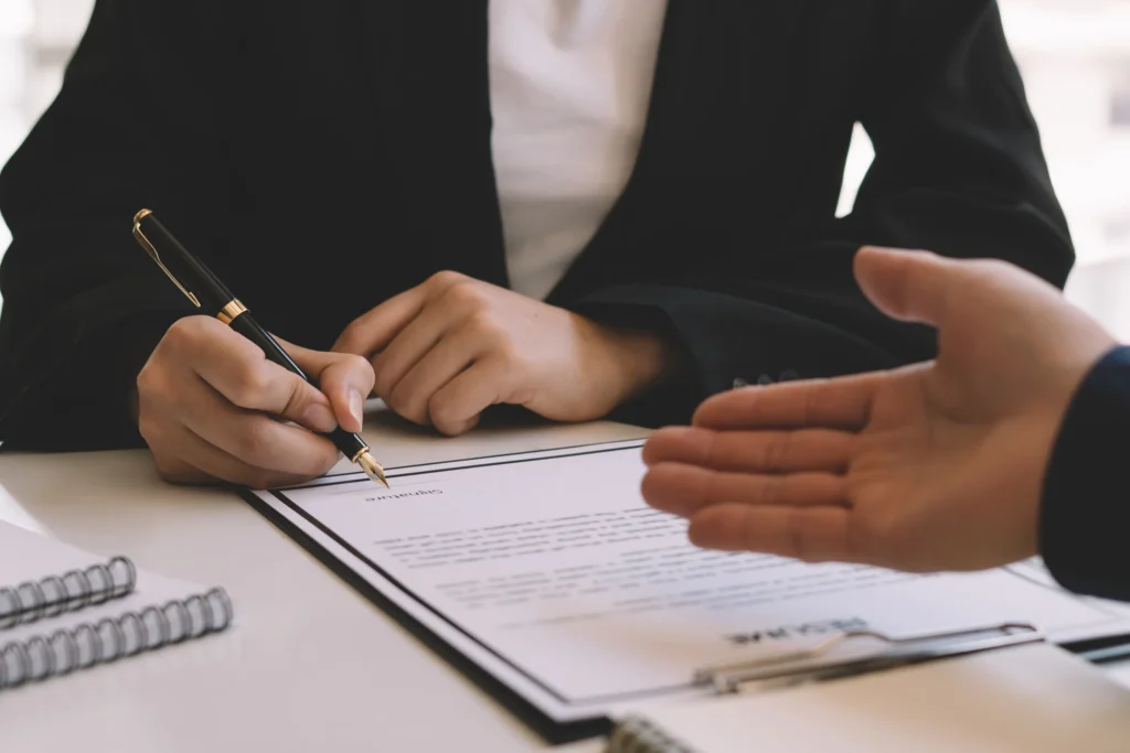 Close-up of a person signing a contract document with a pen while another person gestures across the table, symbolizing legal assistance with contracts in Houston.
