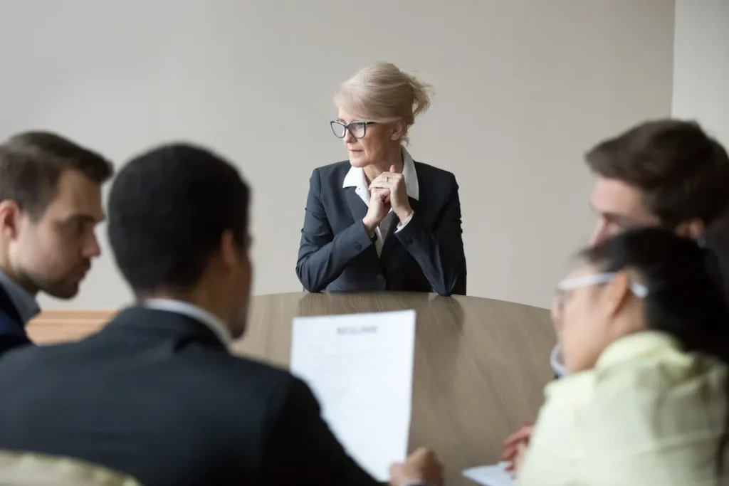 Group of professionals in a meeting, with a serious-looking older woman in a business suit sitting at the head of the table, symbolizing a Houston employment litigation lawyer handling workplace disputes.